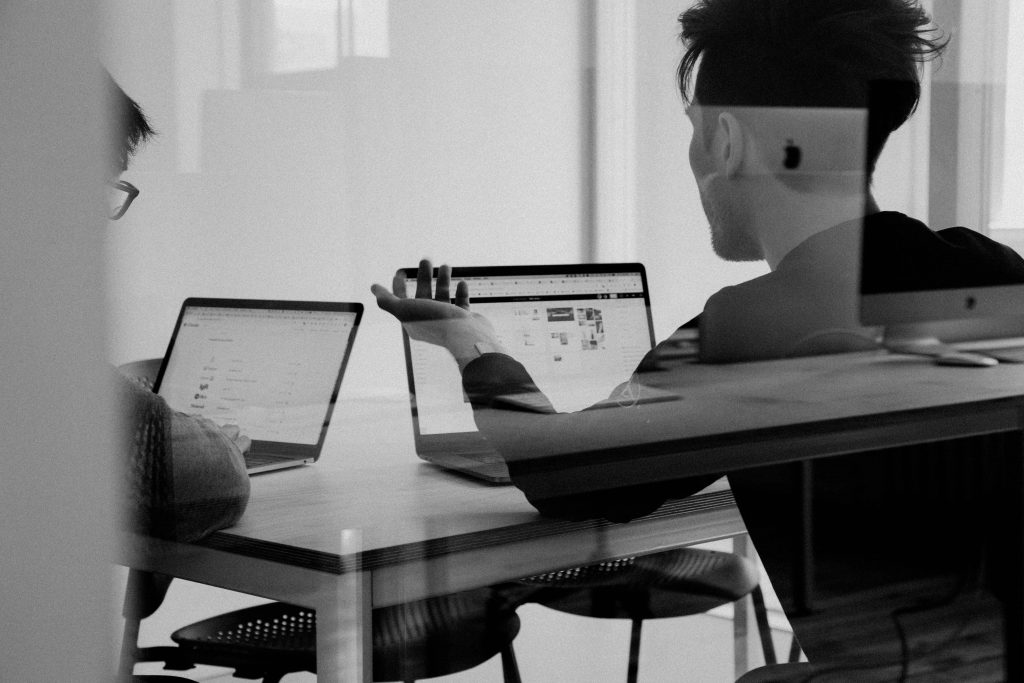 Dois homens sentados em uma mesa, mexendo em computadores. Imagem escolhida para passar ideia de ambiente corporativo