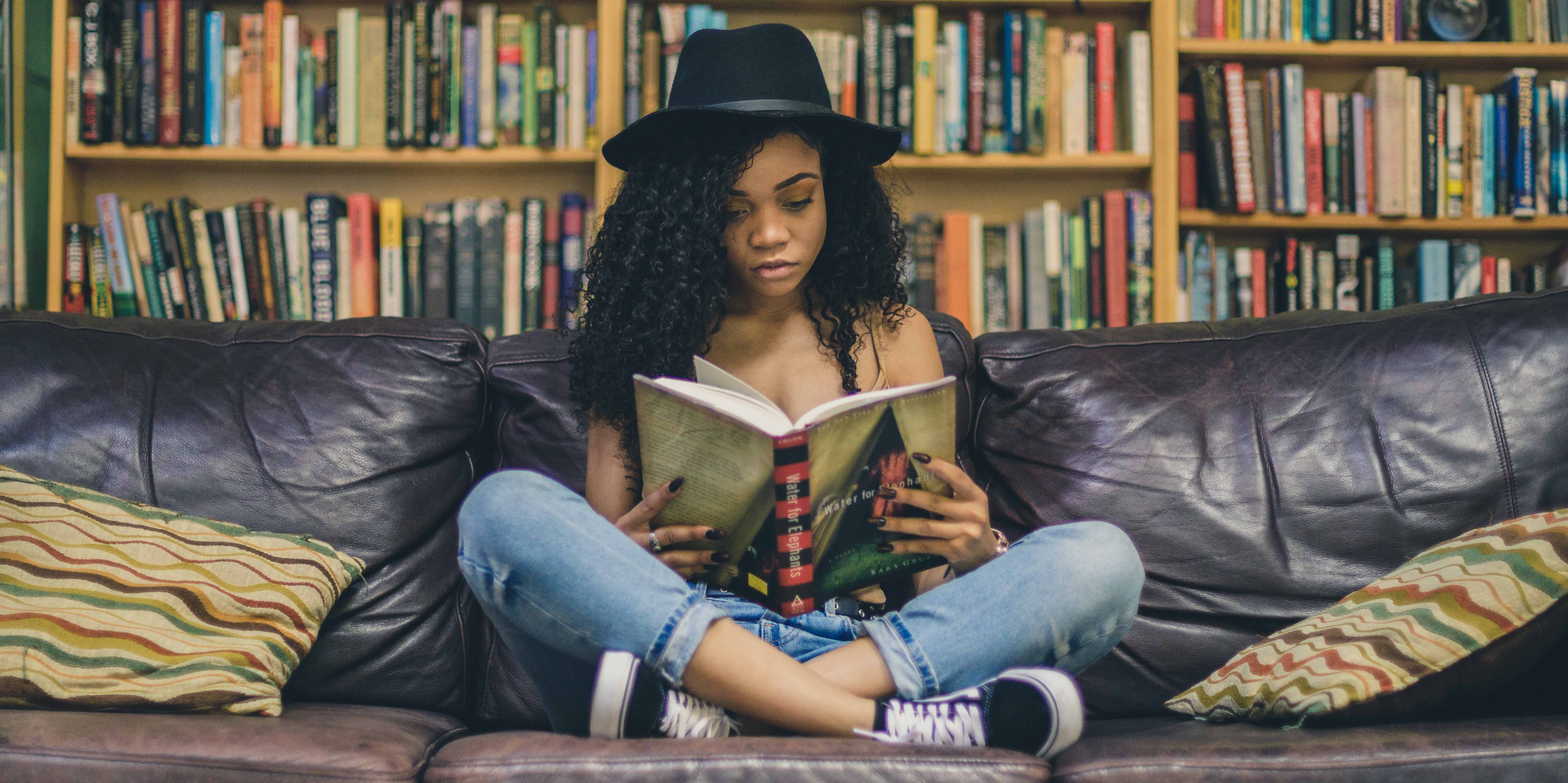 Jovem negra com chapéu preto sentada em um sofa, lendo um livro